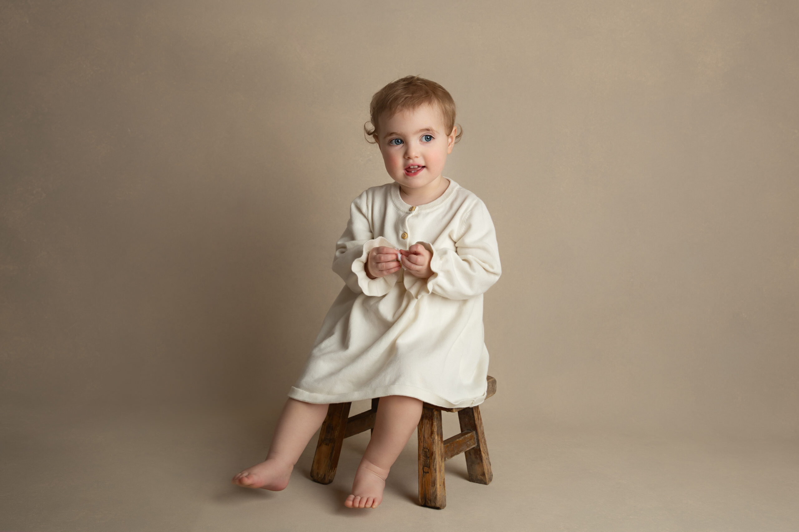 Toddler dressed in a cream long sleeve dress sitting on a small wooden stool.